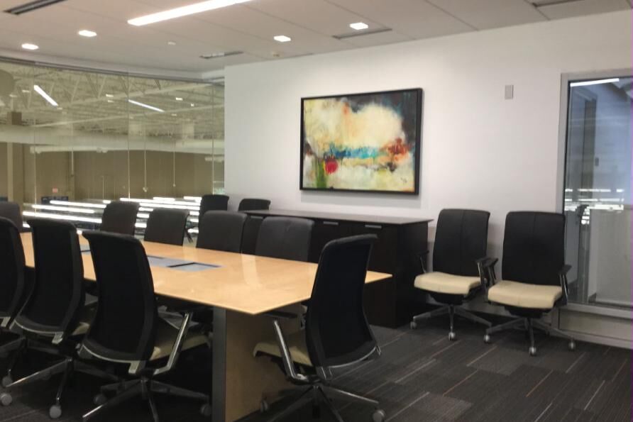 A corporate boardroom: long light-wood table, black task chairs, an abstract painting on a white wall and a glazed partition to the office floor beyond.