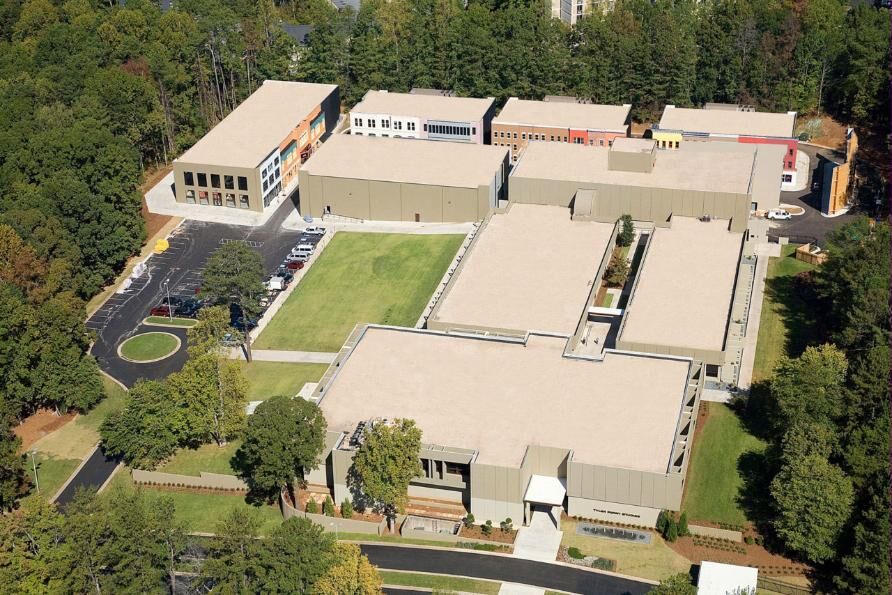Aerial view of a large low-rise studio complex with pale roofs arranged around a green lawn, a parking apron and surrounding woodland.