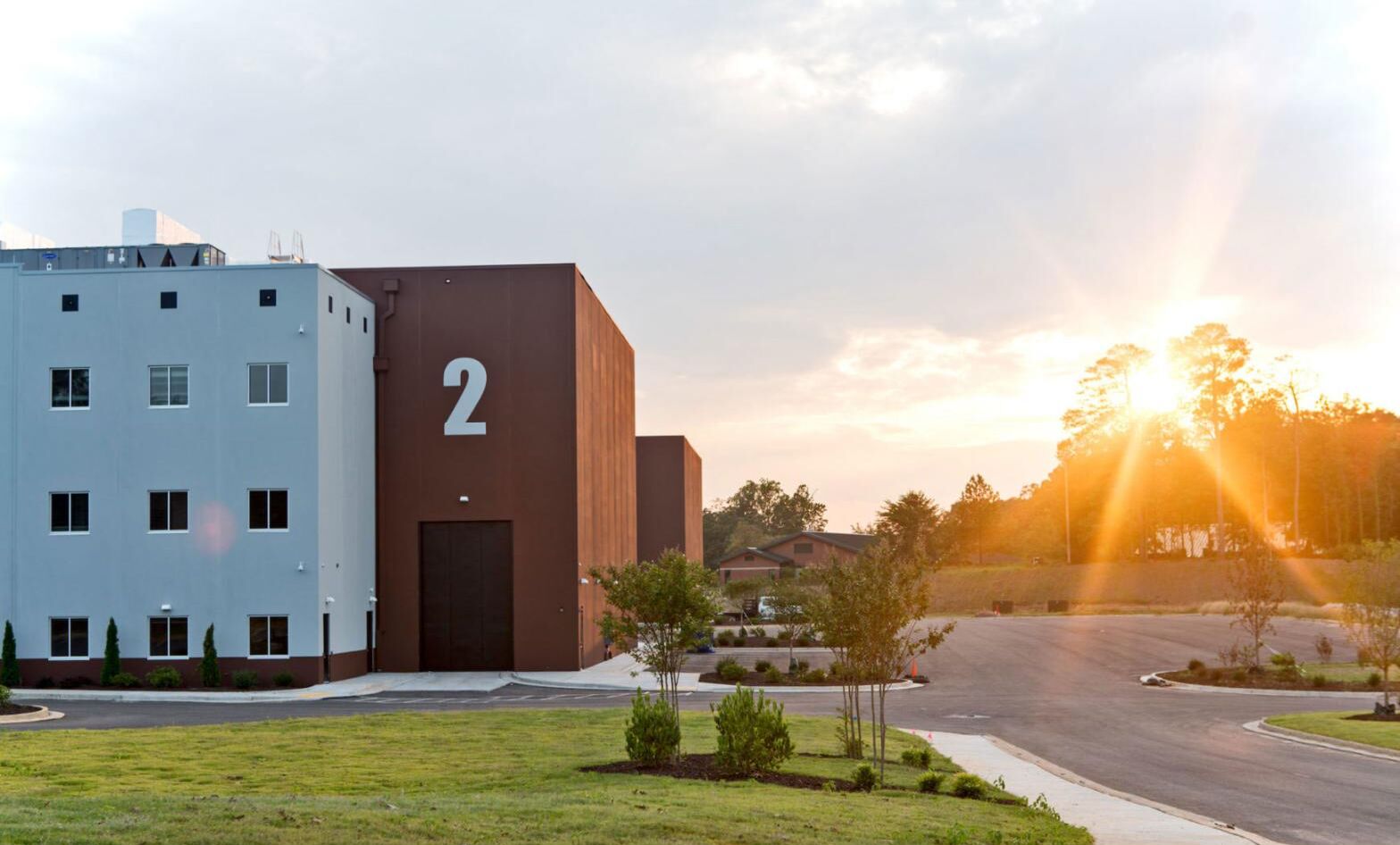 A soundstage marked with a large numeral 2 beside a pale blue office block at golden hour, with landscaped drive and low sun flaring through the treeline.