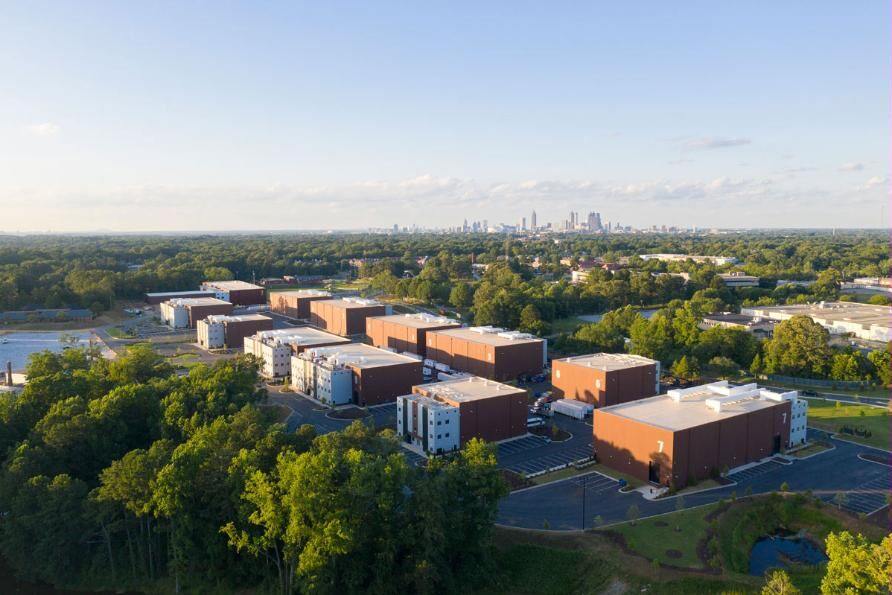 Aerial view of a studio campus: rows of tan and brown soundstage buildings among green treeline, with the Atlanta skyline on the horizon.