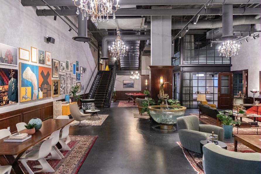 A double-height industrial lobby: polished concrete floor, exposed ductwork, crystal chandeliers, a black steel stair, a stone fountain, mid-century lounge seating and a wall densely hung with colourful art.